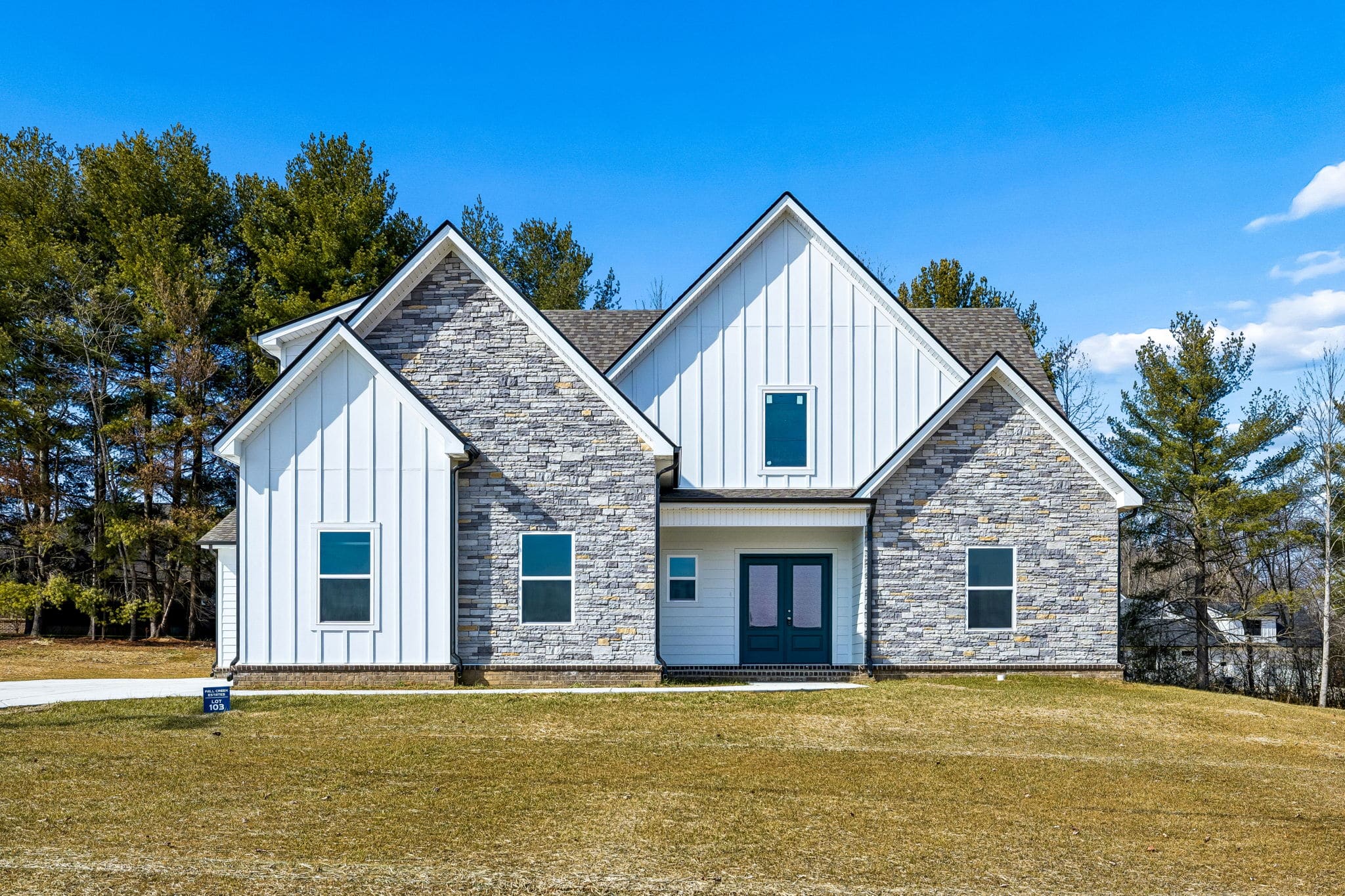 Modern farmhouse with stone and siding exterior, surrounded by trees under a clear blue sky.