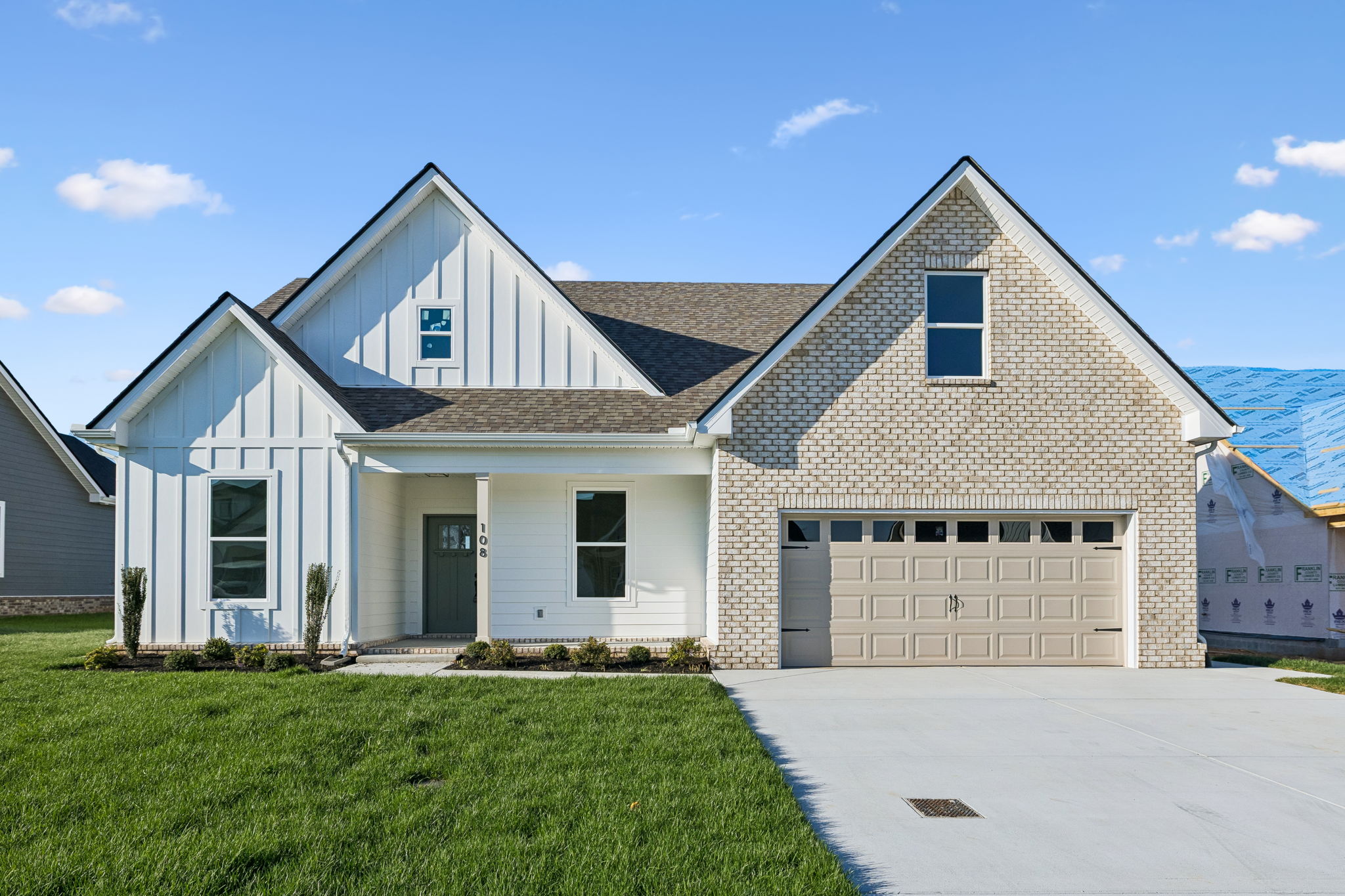 Modern suburban house with white siding, brick facade, two-car garage, and green lawn under blue sky.