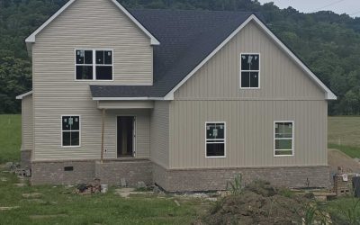 Two-story house under construction on a grass-covered lot, surrounded by trees.