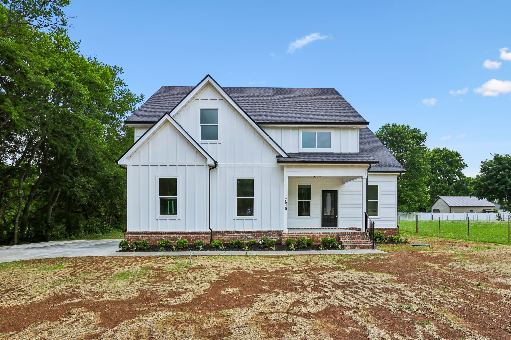 Modern white house with black roof and large windows, surrounded by greenery and blue sky.