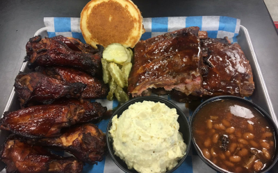 BBQ platter with ribs, chicken wings, cornbread, pickles, mashed potatoes, and baked beans on a tray.