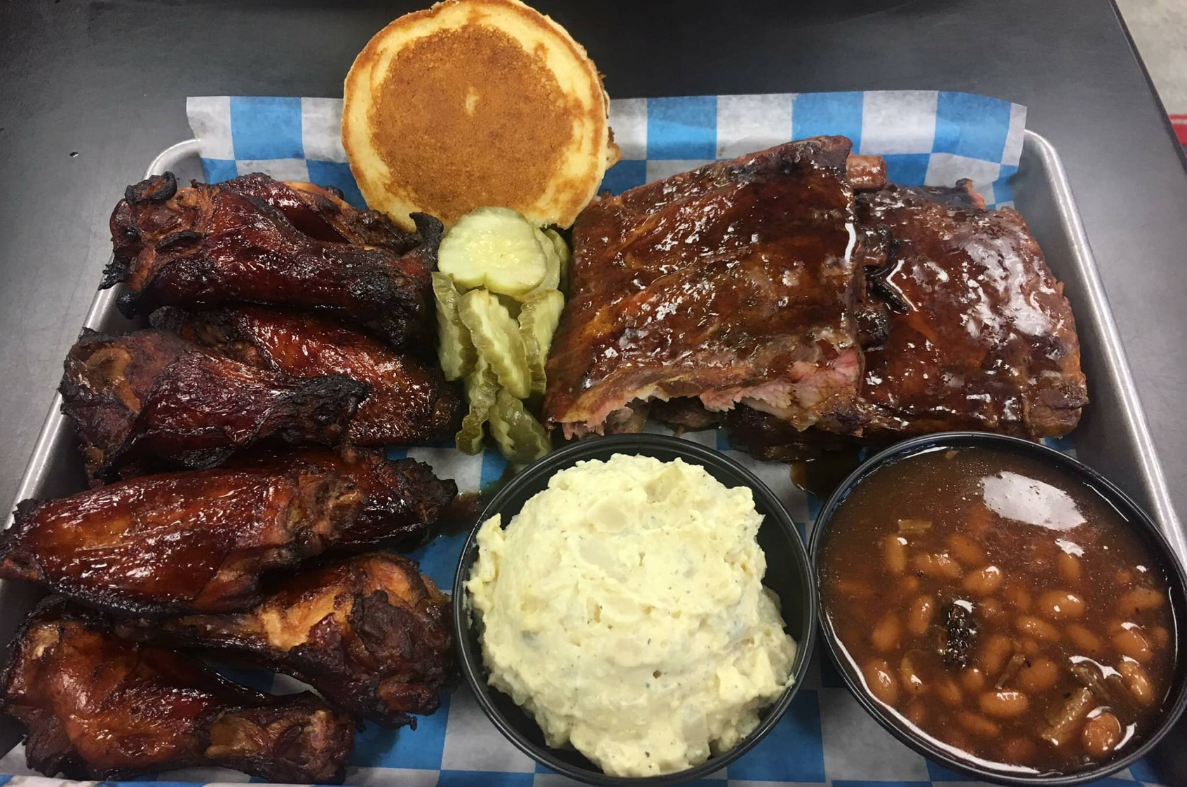 BBQ platter with ribs, chicken wings, cornbread, pickles, mashed potatoes, and baked beans on a tray.